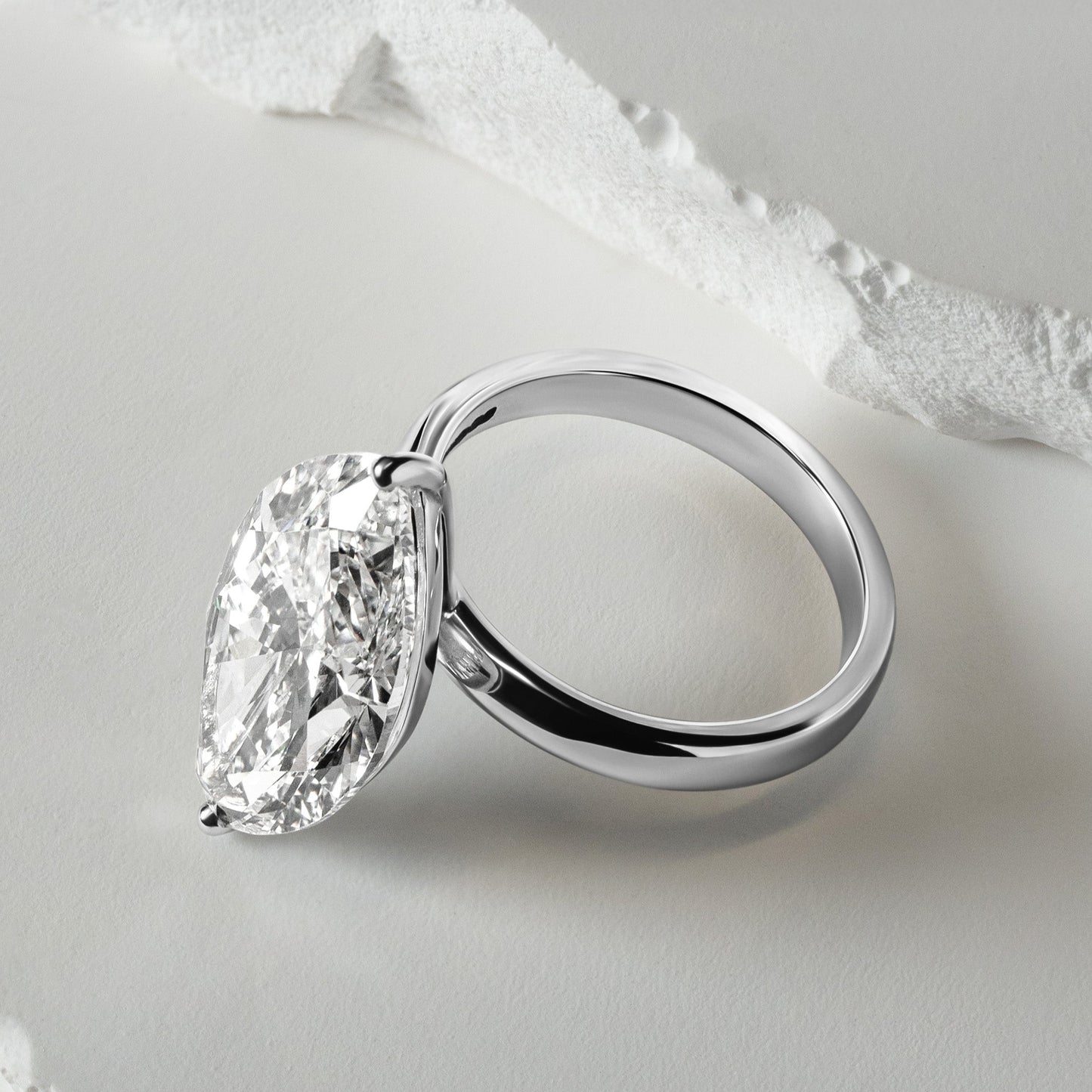 A close-up of a solitaire lab-grown diamond engagement ring with a brilliant oval-cut diamond in a sleek platinum setting, displayed against a textured white background for a refined, modern look.