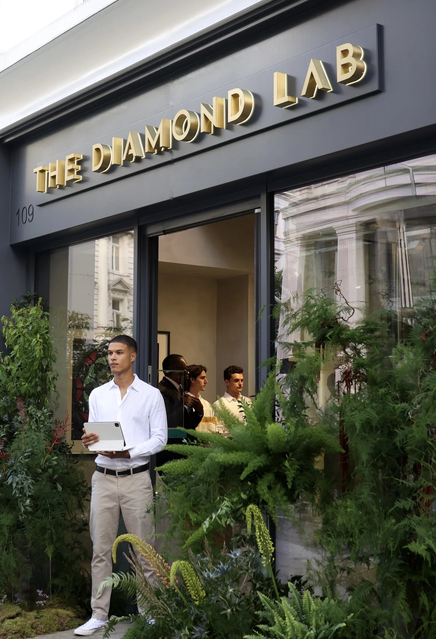 Exterior of 'The Diamond Lab' showroom, featuring sleek gold signage and a modern glass facade surrounded by lush greenery. A man in a white shirt stands at the entrance holding a tablet, while guests mingle inside the brightly lit space.