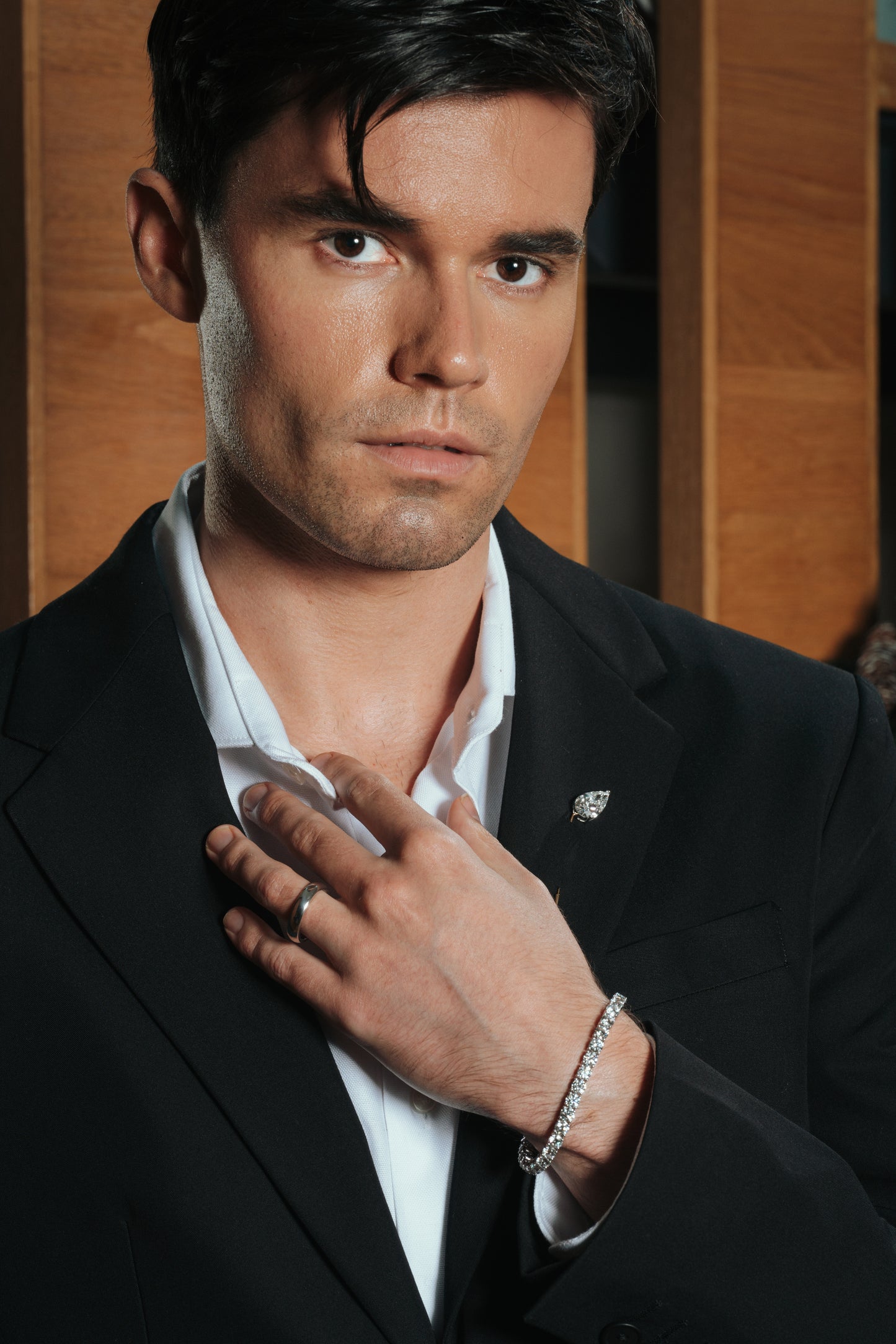 A well-dressed man in a black suit and open white shirt, showcasing a sleek lab-grown diamond bracelet on his wrist and a minimalist silver ring. The elegant setting features a wood-paneled background, highlighting a refined and modern style.
