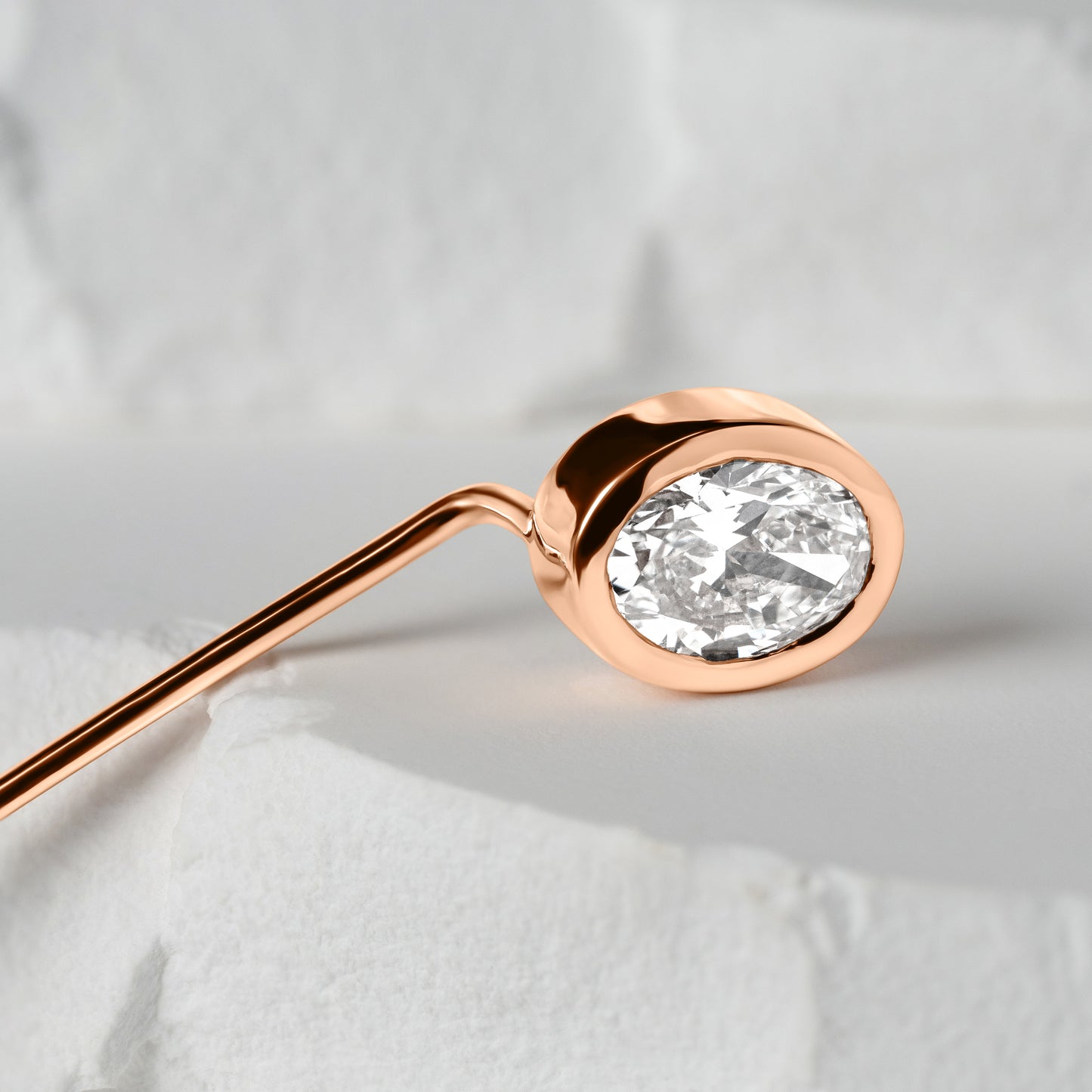 Close-up of an oval-shaped lab-grown diamond set in a polished rose gold lapel pin, resting on a soft white textured surface.