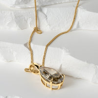 A close-up of a pear-shaped lab-grown yellow diamond pendant in a gold basket setting, resting elegantly on a textured white surface. The pendant is paired with a delicate gold chain, showcasing its intricate craftsmanship from a side angle.