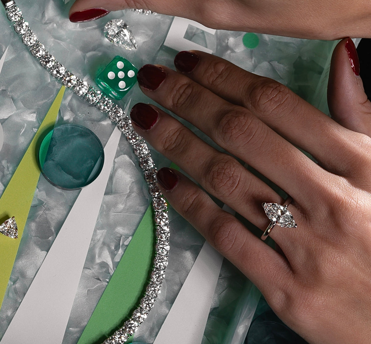 A close-up of a hand with polished red nails wearing a stunning marquise-cut lab-grown diamond ring. The hand rests beside a sparkling diamond tennis bracelet, displayed on a colorful, geometric background with decorative elements.