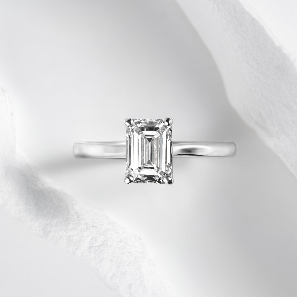 An emerald-cut lab-grown diamond solitaire engagement ring set in a sleek platinum band, displayed against a clean, textured white background for a minimalist and luxurious presentation.