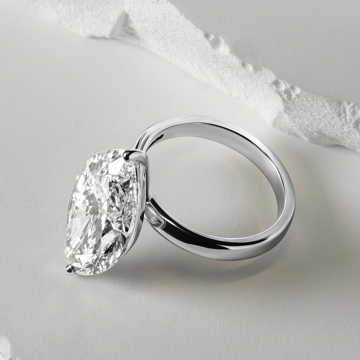 A close-up of a solitaire lab-grown diamond engagement ring with a brilliant oval-cut diamond in a sleek platinum setting, displayed against a textured white background for a refined, modern look.