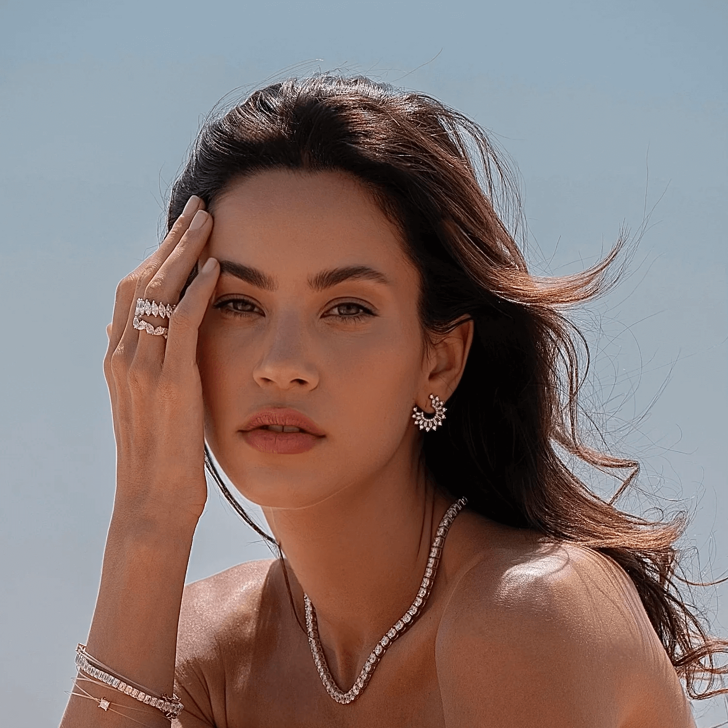 A woman in natural sunlight wearing lab-grown diamond jewelry, including a necklace, earrings, and rings. Her tousled hair and serene expression complement the soft blue sky in the background, creating an effortless and luxurious look.