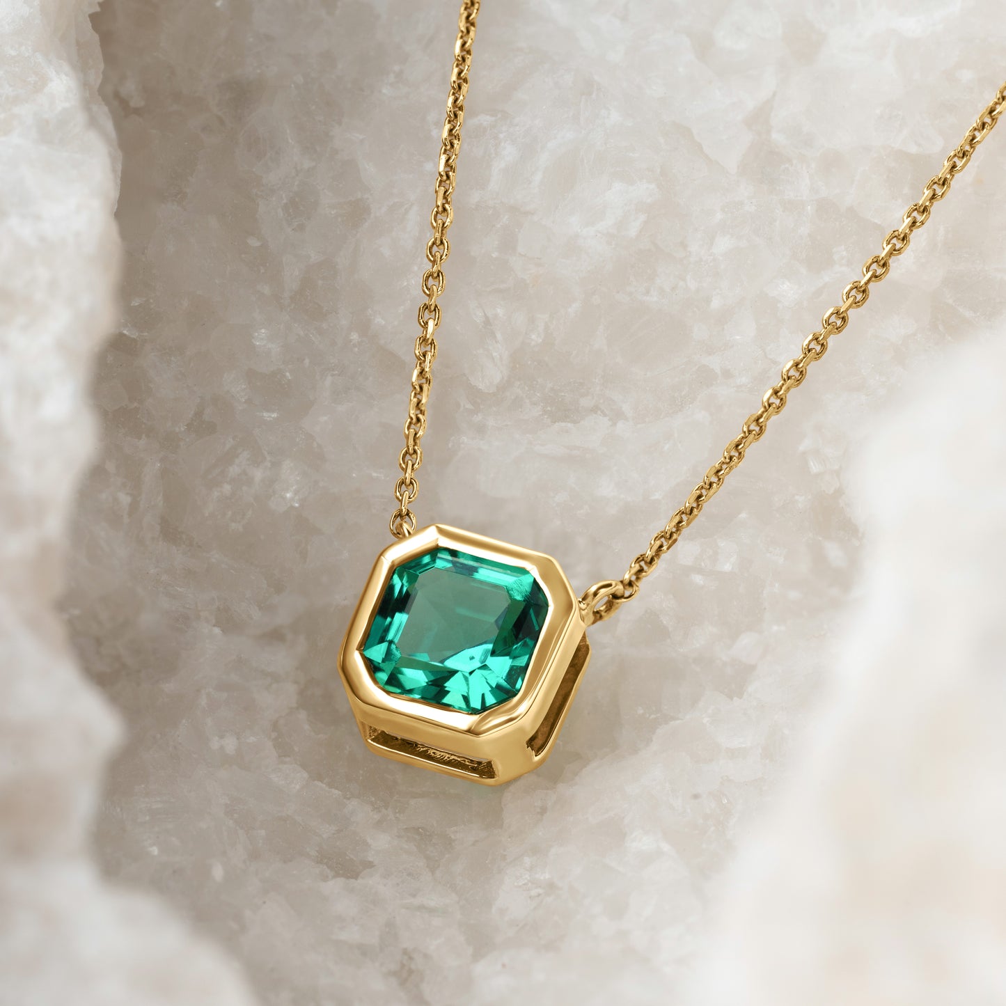 A stunning green lab-grown gemstone pendant in an asscher-cut shape, encased in a sleek gold bezel setting, hanging from a delicate gold chain. The pendant is displayed against a textured white crystalline background.