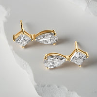 Pair of gold drop earrings featuring pear-shaped and square-cut lab-grown diamonds, displayed on a textured white backdrop with sculptural detailing.
